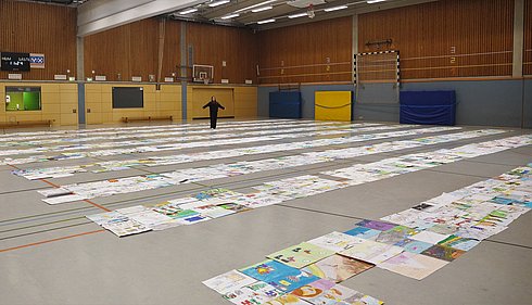 Blick in eine Sporthalle. Der Boden ist mit gemalten Bildern ausgelegt.